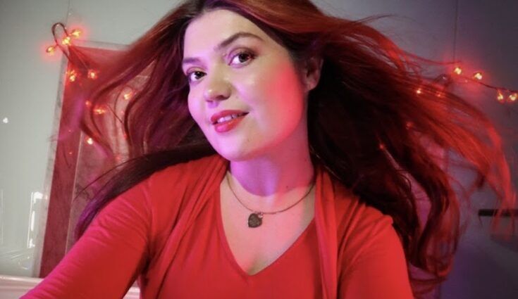 Female with bright red hair and a silver heart-shaoed necklace. She is lit up with red and pink lights shining onto her.